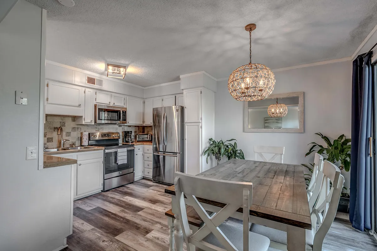 This image presents a dining area with a central table, elegant chandelier, and kitchen appliances visible in the background. The space is set up for family meals, entertaining, and warm gatherings.