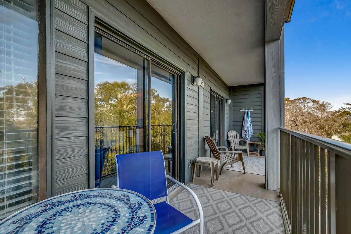 A serene private balcony featuring a mosaic table and wooden chairs set against the balcony railing. The perspective shows a tranquil green space beyond the railing, dotted with mature oaks and flowering hydrangeas, with glimpses of the ocean in the distance. The setting evokes the gentle sea breeze and is ideal for relaxing with a coffee or enjoying the sunset.