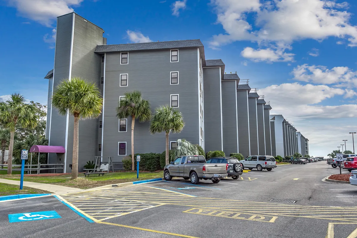 A lively exterior of the resort’s tall buildings, bordered by palm trees and a casual parking area where guests can be seen walking or socializing. The setting conveys a sense of arrival and community within the resort environment.