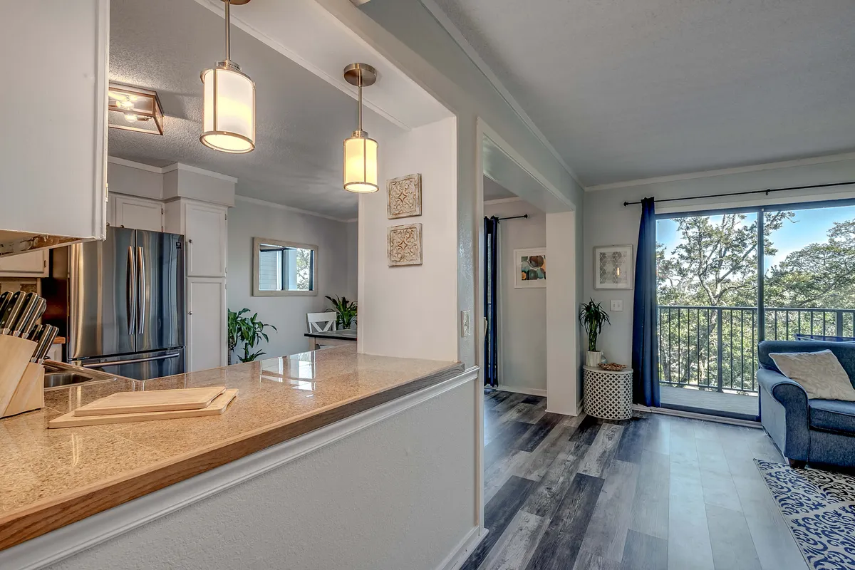 This image highlights an inviting kitchen island that opens into a comfortable living room with a blue couch, and features a visible balcony with partial outdoor views. The setup demonstrates the blend of functional indoor living spaces with easy access to outdoor relaxation, such as enjoying coffee on a private balcony or entertaining guests indoors.
