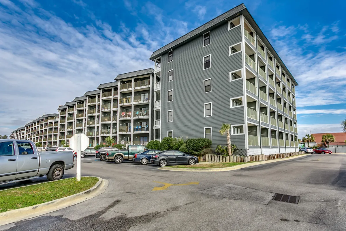 A multi-story resort building with numerous balconies overlooks well-maintained landscaping and palm trees. The parking area is visible, and the scene conveys a sense of arrival and security, with a welcoming vibe typical of a gated resort community.