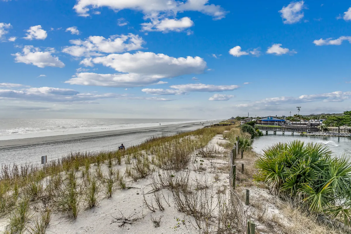 This image captures a sweeping beachfront view with golden sand leading to the ocean, bordered by coastal vegetation and distant resort buildings. The scene is tranquil, with space for families to walk, play, and enjoy the surf.