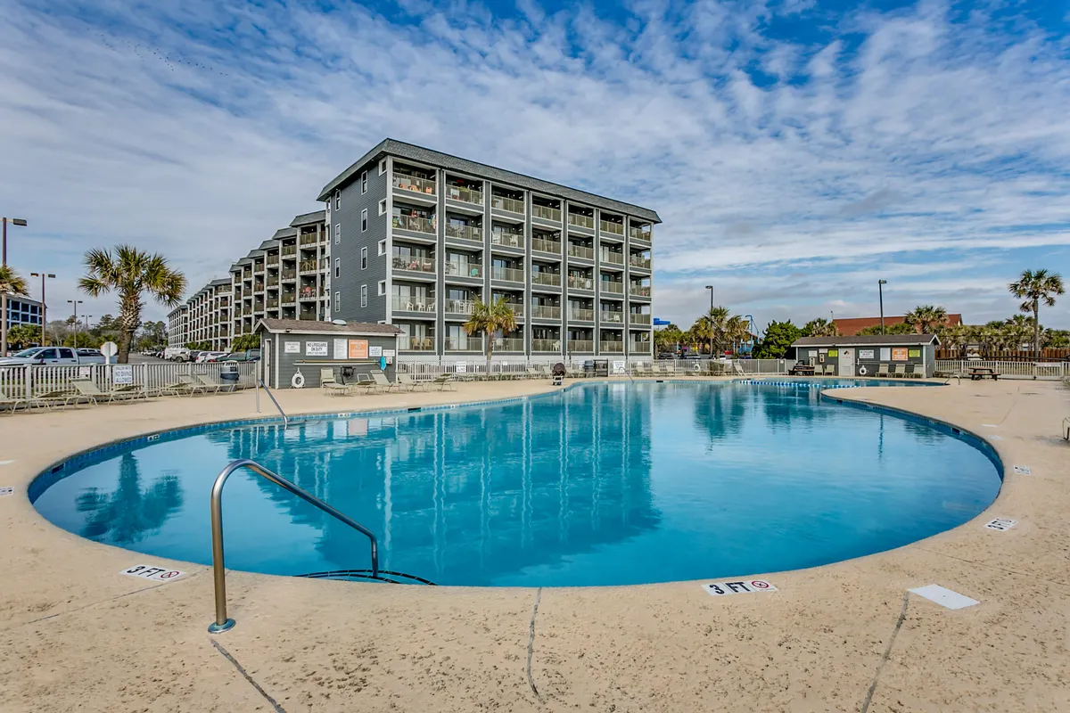 A sparkling swimming pool framed by palm trees and a modern resort building, inviting families to swim, sunbathe, and enjoy the lively resort atmosphere. Lounge chairs and open spaces suggest group recreation and relaxation.
