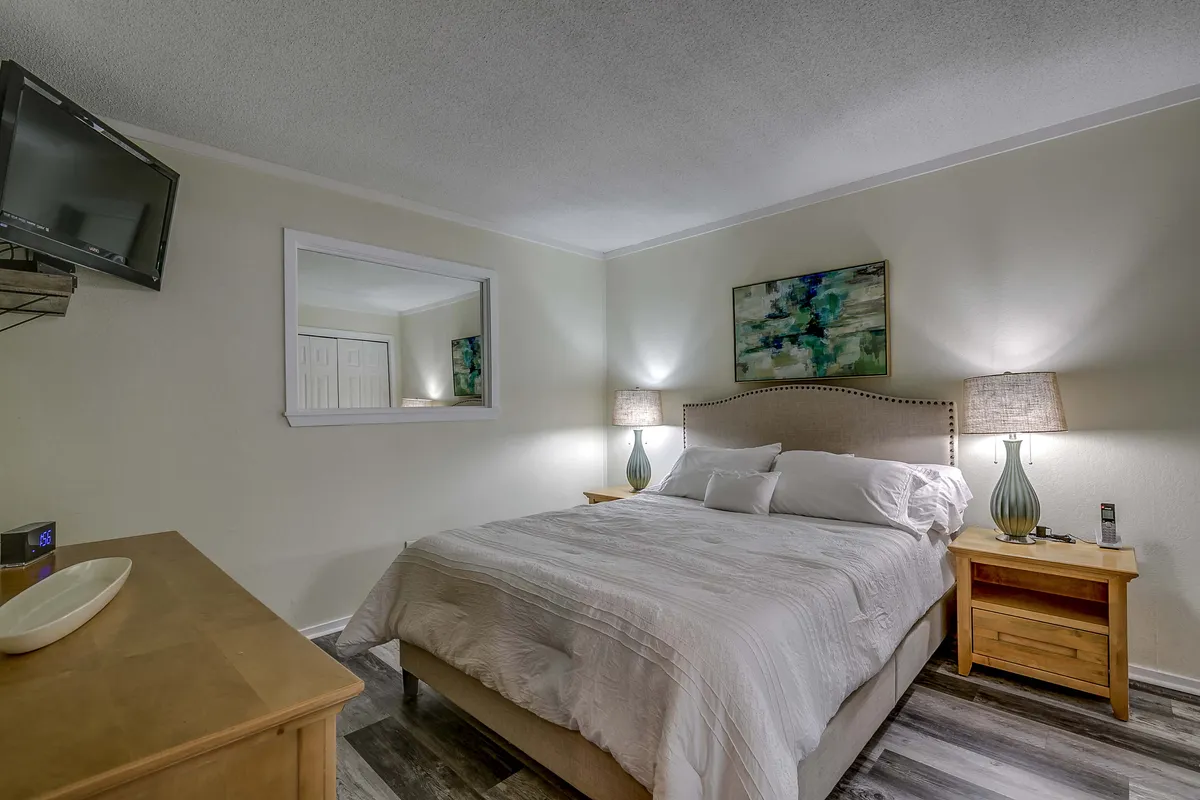 This image captures a tranquil master bedroom setting, featuring a cozy queen-sized bed with plush linens, bedside nightstands and lamps, tasteful artwork above the bed, and a wall-mounted television. The soft lighting and calm palette create a peaceful and inviting atmosphere.