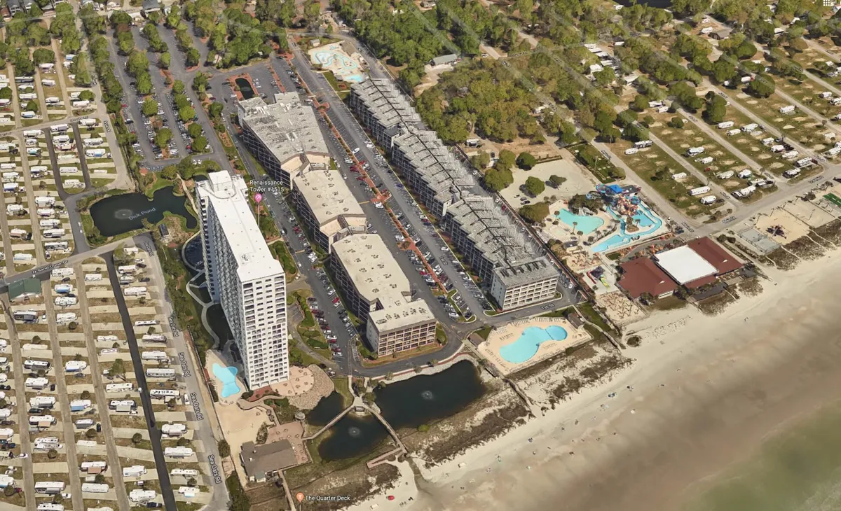 An exterior scene showing a tall white hotel building with rows of balconies, surrounded by landscaped recreational areas, including swimming pools and camping sites. The foreground hints at communal spaces ideal for family gatherings, outdoor dining, or BBQs, with picnic areas visible below the balconies.