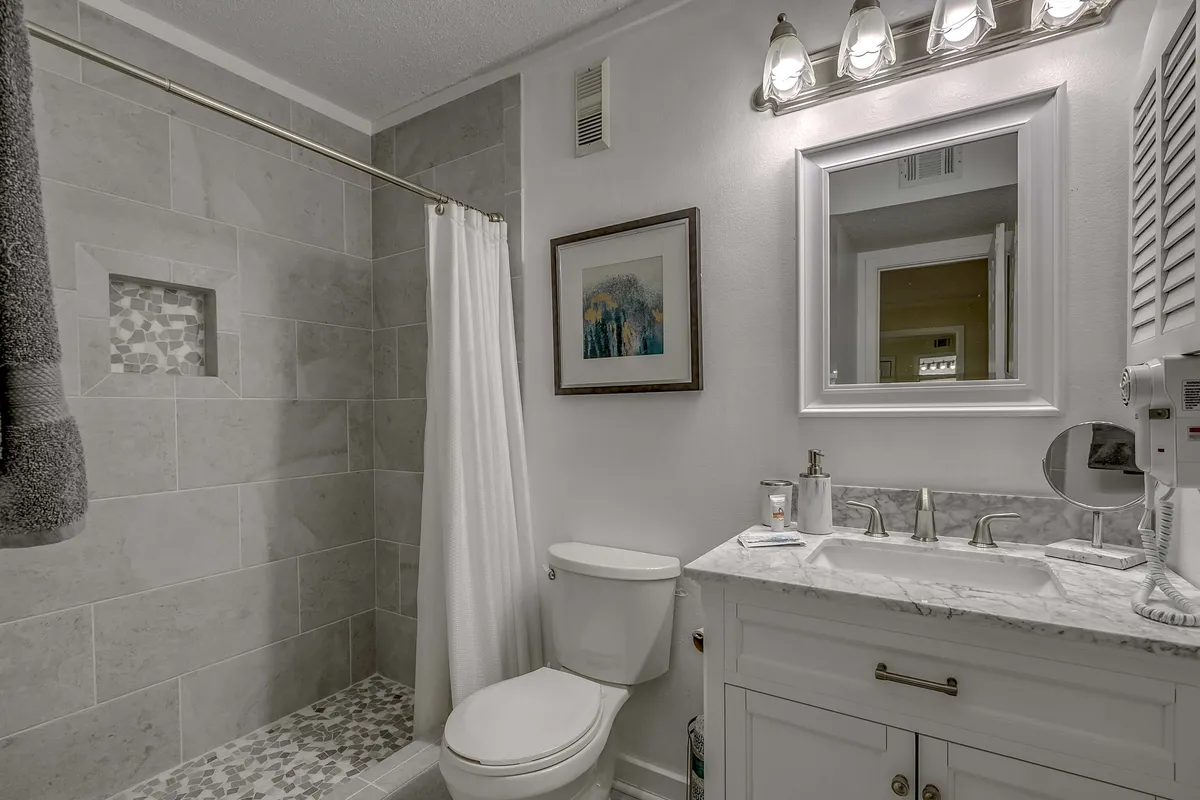 A bright and modern bathroom scene showcasing a sleek shower area, a vanity with Carrara marble-style countertop, a large mirror, and framed artwork. Plush white towels are neatly arranged, contributing to the spa-inspired feel.