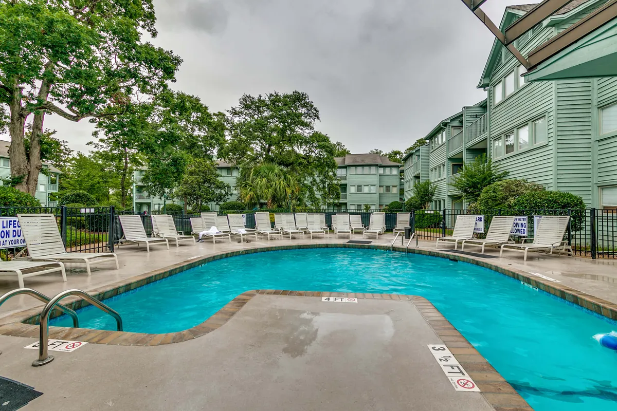 This image showcases the resort's main swimming pool surrounded by comfortable lounge chairs, mature trees, and adjacent apartment buildings. The tranquil pool area is sun-dappled and inviting, perfect for relaxation or socializing with neighbors. The setting evokes a sense of calm and leisurely living, ideal for retirees and snowbirds seeking a stress-free environment.