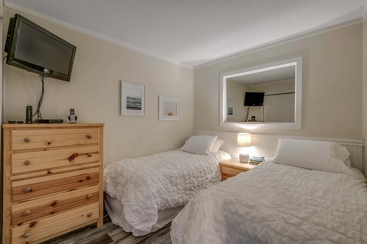 A welcoming second bedroom with two twin beds arranged side by side, each with inviting bedding and plush pillows. The room also features a wooden dresser, decorative framed pictures, a mirror, and a TV/DVD setup on a side table, making it ideal for kids or friends.