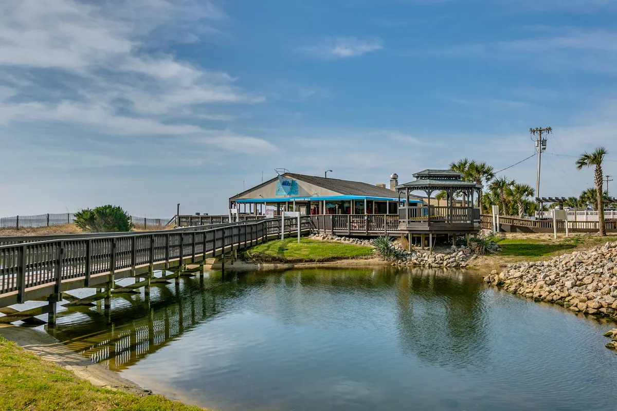 A tranquil exterior scene featuring a wooden bridge leading to a gazebo, with the main resort building in the background. The area evokes leisurely family strolls, exploration, and outdoor bonding moments.