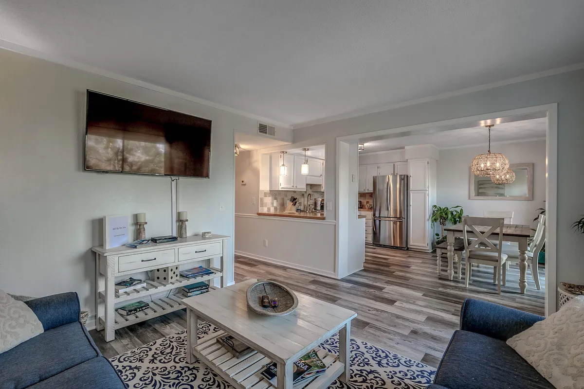 The image showcases a bright, inviting open-plan living area featuring a plush seating area, a spacious dining table set for family meals, modern kitchen appliances, and a stylish decorative chandelier. The scene exudes warmth and comfort, suggesting a perfect space for family gatherings, casual dining, and relaxing together after a day at the beach.