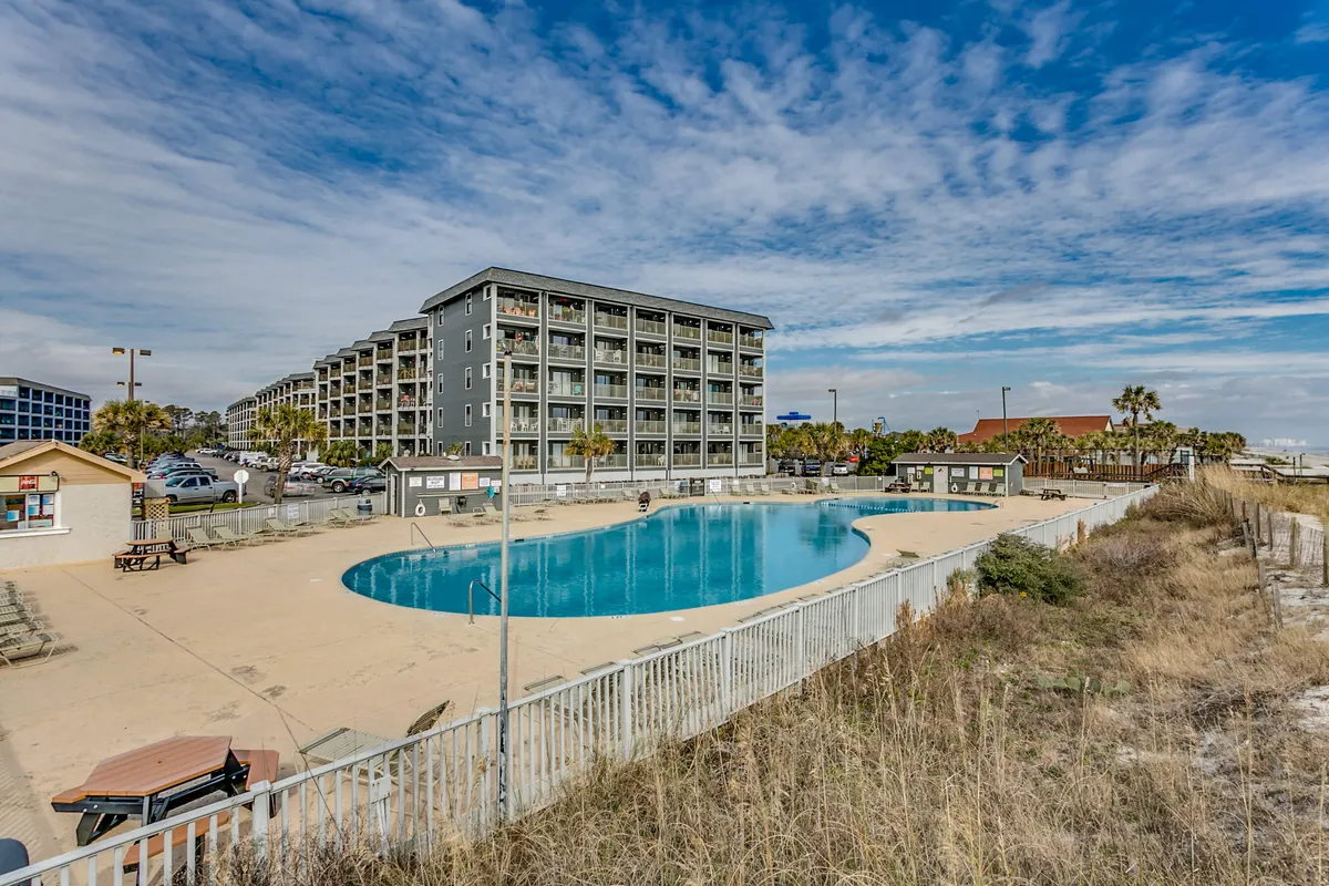 A vibrant swimming pool scene framed by palm trees and the main resort building, featuring areas for lounging, social gatherings, and enjoying outdoor amenities. The landscape is welcoming and suggests both activity and relaxation options for larger groups.
